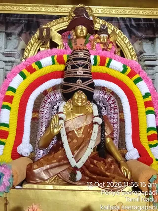 A golden murti of a seated goddess, identified by the page as Sri Kamakshi Amman, decorated for Navarathri at the Periya Seeragapadi temple in Tamil Nadu. The figure wears a rust-brown and gold-bordered silk sari, a long gold necklace and a white flower garland, with a black, gold-banded tall crown above her head. Her right hand is raised in abhaya mudra. Behind her an arch of woven garlands in pink, white, red, yellow and green frames a larger golden shrine arch, with a smaller golden deity figure visible above. Pink flowers and orange cushions surround the base, and a photo overlay shows the date and location.