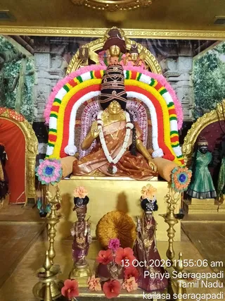 A golden murti of a seated goddess, Kamakshi Amman, is decorated for alankaram in a temple shrine. She sits on a gold pedestal with a raised right hand in abhaya mudra, wearing a brown and gold silk sari, layered gold necklaces and a tall dark crown with gold ornaments. A large arch of pink, white, yellow, red and green flower garlands frames her, with white jasmine garlands draped on her shoulders. Two smaller dark-faced attendant figures in brown silk stand at her base, one holding a staff, surrounded by red hibiscus and orange flowers. Tall brass oil lamps flank the pedestal, and a black-faced figure in a green sari stands in a side niche. No physical people are present.