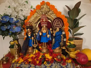 A home shrine arranged for a puja with three golden murti of deities in the centre, each wearing blue garments and rudraksha malas, the central figure with a red and white patterned crown and raised hands. Behind them is an arch of golden metalwork encircled by orange marigold garlands. Red and yellow flower petals are scattered across a blue sequined cloth in front, with a golden trishul, a brass oil lamp, brass pots of yellow flowers, bananas, a pomegranate and a mango on the right, and blue and white flowers on the left. No people are visible.