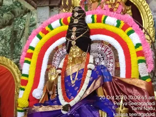A golden deity murti seated on a throne is the central subject, adorned with a tall black hair crown fitted with gold ornaments and round medallions. It wears a blue sari with gold border, a long white and red garland, gold necklaces and bangles, and holds one hand raised in blessing. Behind it an arch of yellow, red, white and green floral garlands framed by pink flowers surrounds the shrine, with a silver-patterned backdrop and a brass lamp visible at lower right. A timestamp reads 20 Oct 2026 10:09:45 am.