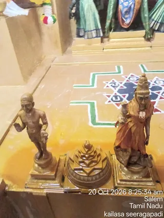 A photograph of a shrine floor with three bronze-coloured figures on a golden platform before a rangoli-style geometric kolam of green, white, red and dark blue lines. On the left stands a small bare-chested male statue in a loincloth with one hand raised. In the centre sits a stepped conical sri yantra-like pedestal. On the right stands a female deity murti in an orange-red garment with a tall crown, holding a small object in one hand. Behind them, painted figures in green skirts and blue shields appear on a shrine wall. A timestamp reads 20 Oct 2026 8:25:34 am, Salem, Tamil Nadu, kailasa seeragapadi.