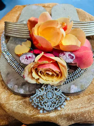 Close-up photo of a pair of ornate golden-brown sandals or padukas resting on a gold brocade cloth, decorated with orange and peach roses, scattered rose petals, two small pinkish-red stones or sweets, and a sparkling crystal necklace with a large pendant set with white stones and dangling pearl drops. No people are visible.