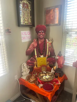 A corner altar room with a seated figure in a red turban and red robes, sitting cross-legged on an orange cloth-covered platform. The figure wears a rudraksha mala and white flower garland, holds a golden trident, and raises one hand in blessing. A small pink paper sign reads KAILASA SAN DIEGO on the wall. Around the figure are a white marble Ganesha statue, a gold yantra plate, brass puja tray with white flowers, a bowl of red powder, a small oil lamp, and a dark statue wearing a red cloth. A framed gilt Tirupati-style deity image hangs above. Because the figure is a ceremonial seated presentation with a turban, rudraksha mala and trident, the appearance cues are distinctive; the person is described as a seated devotee-style presentation within the altar.