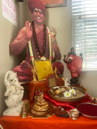 A home shrine photographed indoors beside a window with white blinds. Dominating the upper left is a life-size cardboard cutout of a smiling man in a red turban, rudraksha mala, white flower garland and saffron-red shawl, holding a trident and raising one hand. The cutout is a depiction, not a physical person. Below it, an orange cloth covers a table holding a white marble Ganesha seated on a lotus, a golden yantra plate framed in gold, small gold deity figurines, a red-and-gold dotted deity murti, a gold Meru-shaped object ringed with rudraksha, a brass plate with white chrysanthemums and brass cups, a silver kalasha-style cup, a tray of red kumkum and a rudraksha mala. A paper sign on the left wall reads KAILASA SAN DIEGO.
