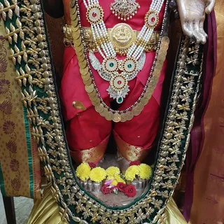 Close-up photograph of a deity murti dressed in a red garment with gold and pearl ornaments. The figure wears a layered necklace of gold coins, a broad gold waist belt with a central medallion, and multi-strand pearl and red-green stone jewellery. Yellow chrysanthemums and red roses lie at the feet on a silver base, and an ornate green and gold-embroidered cloth frames the lower figure. Only a carved hand is visible at upper right. No physical people are shown.