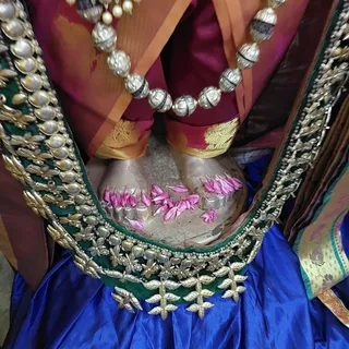 Close-up of a silver-toned deity murti seen from above, its lower body draped in a magenta and orange silk garment with gold border and a deep blue satin skirt trimmed with green and gold embroidered lace. A long necklace of silver beads and pearls hangs across the garment. Pink petals are scattered over the murti's base and a small silver flower-shaped setting near the bottom, consistent with a puja offering. No people are visible.