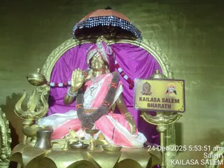 A golden statue seated on a gilded lotus pedestal in front of a purple cloth backdrop and an ornate gold arch, its right hand raised in blessing. The figure wears a pink and white garment, a pink and white turban with a crescent-like ornament, a garland of red and white flowers, and a dark brown flower garland draped across the chest. A gold trident stands at the left, with a brass vessel, a bowl, a banana, a small brass dish and a red-and-orange canopy above. A printed sign on the right shows a small portrait labelled KAILASA SALEM BHARATH. The statue is a deity murti in an ashram shrine; a timestamp reads 24-Dec-2025 5:53:51 am Salem.