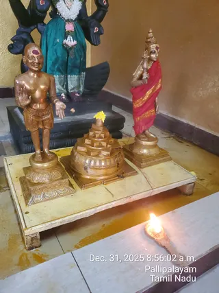 Indoor shrine photograph showing three bronze-coloured metal figures on a low cream wooden platform. On the left stands a bare-chested figure with a red tilak on the forehead wearing a short dhoti, raising one hand in blessing. In the centre sits a tiered sri yantra-style metal base topped with a yellow flower. On the right stands a figure in a red shawl with gold border, a tall crown and a red mark on the forehead, holding a small ritual object. Behind them a dark deity murti in a teal sari with flower garland is partly visible. A small oil lamp burns at the front edge of the platform, and the floor is wet with spilled liquid. A timestamp overlay reads Dec. 31, 2025 8:56:02 a.m. Pallipalayam Tamil Nadu.