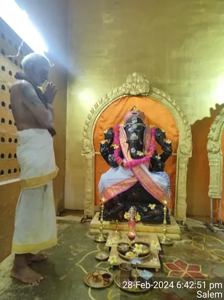 Photograph inside a temple shrine. A bare-chested elderly man with white hair, wearing a white dhoti with a gold border, stands at left with palms pressed together in namaskar, facing a black stone Ganesha murti seated in an arched golden niche with an orange backdrop. The murti is garlanded with pink and white flower strands, draped with a pink and gold silk cloth, and decorated with yellow and white flowers. Brass oil lamps and offerings in brass bowls with flower petals sit on a wooden platform in front, with a kalasha-like vessel and a lit lamp. A kolam design decorates the floor. The man is a devotee at the puja, with no identifiable distinctive a person cues.