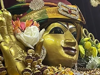 Close-up photograph of a golden deity murti with a glossy gold face, painted eyes and a red-and-green turban decorated with a jewelled crest and a red stone. A white rose and orange and pink flowers rest against the figure, with a hand raised beside the face. Rudraksha malas and gold garlands hang at the bottom of the frame, and yellow flowers appear at the right. No physical people are visible.