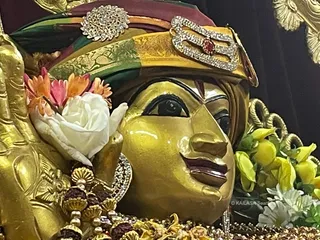 Close-up photograph of a gold-painted deity murti with a serene face, dark almond eyes and a red-brown lip, wearing a multicoloured turban with gold brocade and a jewelled emblem set with a red stone on its front. White and pink-orange flowers are tucked at its side, with yellow blossoms and white flowers in the background, and rudraksha beads and a gold garland at the lower edge. The setting is a temple shrine with a dark backdrop and a gold-framed element at the top right.