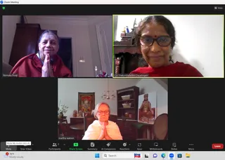 A screenshot of a Zoom video call with three participant tiles. Top left, a woman in a dark red shawl with hands pressed in anjali mudra in a wood-panelled room. Top right, a smiling woman in a red top and glasses in front of a shelf with small brass figures. Bottom centre, an older woman with glasses in a white garment, hands in anjali mudra, seated in a dining room. Behind her hangs a framed painting of a deity and a portrait of a seated saffron-robed figure with long hair on a red chair, appearance uncertain. Zoom toolbar labels and a weather widget are visible at the bottom.