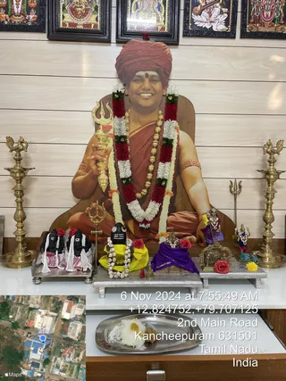 Photo of a home shrine. A large life-size cutout print of a smiling man with a saffron-orange turban and red-orange robe, a vibhuti mark on his forehead, a rudraksha-style gold bead necklace and a garland of white and red flowers, holding a golden trident-topped staff, sits as the centrepiece against a wooden wall. Brass oil lamps flank the altar. Before it stand two black Shiva lingas dressed in white cloth and red flowers, and a black linga with a white bead garland on a yellow cloth. A purple cloth covers a small deity murti, a metal shrine holds another figure, and a steel plate holds white and yellow offerings. Framed deity paintings hang above, and a timestamp and map overlay sit at the bottom right.
