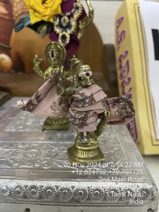 A small brass deity murti stands on a pedestal in the foreground, draped in a pink fabric with a brown floral print and blue piping. The figure has a red tilak mark on the forehead and a raised trident-like tool held in one hand. Behind it, a second brass figure with multiple raised arms stands on a base, next to a bright yellow flower arrangement and a maroon cloth with a jeweled ornament. A yellow box with pink lettering sits at the right edge, and a silver-toned, embossed metal tray with floral and circular patterns is in the lower foreground. A timestamp overlay reads 20 Nov 2024 at 7:14:22 AM with a location of 2nd Main Road, Kancheepuram 631501, Tamil Nadu, India.