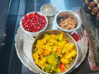 A top-down photo of a steel offering plate on a dark stone or metal surface. The main steel bowl holds yellow pasta with broccoli florets, carrot pieces and red bell pepper. A bowl of pomegranate arils sits to the upper left, a bowl of whole almonds to the upper right, and a small steel cup of water sits at the top centre. A red flower petal lies beside the plate. At the upper right edge, dark rudraksha beads are strung on gold-tipped stands. No people are visible.