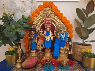 A home shrine photograph showing three brass deity murtis standing before a gilded arched backdrop framed with orange marigold garlands. The central figure wears a tall red-and-white striped crown, a white flower garland and a rudraksha mala, and holds a trident-like staff. The figures on either side wear blue cloth, one with a tall crown of pearls and a long black braid, the other with a patterned headdress and a rudraksha mala. White flowers, potted plants, a brass bell, a red mango-shaped offering, red rose petals, and a small brass Ganesha figure with a blue-and-green bead garland are arranged on a blue and gold cloth in front. No people are visible.