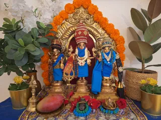 A decorated home shrine with three bronze murtis standing before a gold arch framed by orange marigold garlands. The central figure wears a tall striped crown, a multi-strand white and pearl necklace and a red sash, and holds a trident-like implement. The two flanking figures wear blue cloth, beaded blue necklaces and crowns. Red roses and a red-and-blue garland surround a small brass Ganesha figure and a dark stone linga-like object on a lotus-patterned cloth. A brass bell and a large pink stone sit at the front, with potted plants and white flowers on either side. No people are visible.