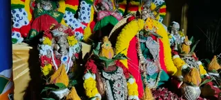 A photograph of a decorated shrine at night showing several deity murti adorned with yellow and red marigold garlands and silver ornaments. Figures in green, red and gold garments and silver crowns stand on a draped altar. Brass kalashas topped with coconut-fibre tufts and green leaves sit at the lower corners, and red flowers are scattered across the base. No people are visible.
