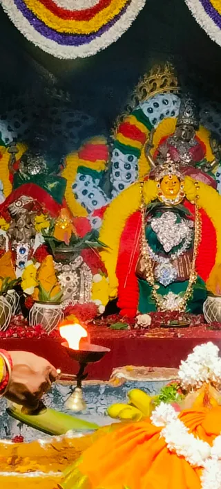A photograph of a decorated goddess murti in a shrine, lit by a brass oil lamp (diya) held at lower left by a hand. The deity has a golden face, a silver crown and a heart-shaped silver chest ornament, wearing a red and green sari draped with yellow and red garlands. Behind her, a second silver-adorned figure and white and yellow flower garlands hang across the altar. Yellow bananas, a fruit offering and a lit lamp sit on the altar in front, with an orange-clad arm and white flower garland at the lower right. Festive pennant-like garlands decorate the top. No people are visible in the altar setting beyond the hand holding the lamp.