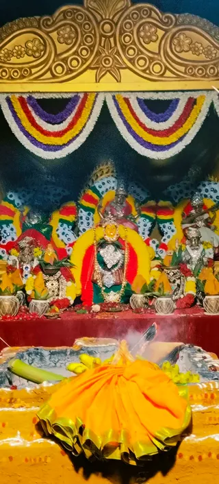 A photograph of a shrine decorated with garlands in red, yellow, purple and white, with a gilded arch above. Silver and gold-covered deity murtis stand at the centre and sides, one central figure draped in a red and yellow shawl and wearing a gold crown and necklace. Brass and silver kalashas with mango leaves and marigold garlands flank the altar. In the foreground a yellow-and-gold cloth covers a fire pit where smoke rises and green banana stalks are placed. No people are visible.