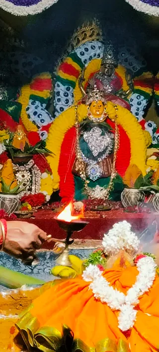 A photograph of a deity murti adorned with yellow and red marigold garlands, a silver heart-shaped chest ornament and layered silver necklaces, seated before a dark backdrop with a crown-like silver halo. A brass oil lamp with a bright flame sits in front, and a hand holds a stick toward it. In the lower foreground, a figure wearing an orange garment and white jasmine garland is partly visible, with smoke rising. Potted plants and flower offerings flank the murti.
