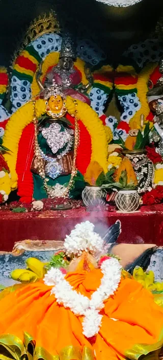 A photograph of a decorated deity murti of a goddess, seated on a red-draped altar framed by yellow and red marigold garlands and blue-dotted cloth backdrops. The murti wears a silver crown, a silver heart-shaped chest ornament, a beaded gold necklace and a green sari with a red border. Two silver vessels with green leaves stand beside it, and red flowers and a banana bunch are arranged on the altar. In the foreground a figure in an orange garment wears a white jasmine garland, with incense smoke rising.