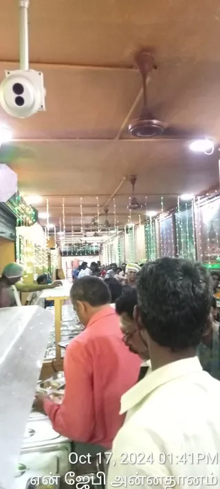 A photograph taken inside a crowded, brightly lit hall during an annadanam food service. In the foreground a man in a pink-red shirt stands at a steel serving counter with food trays and containers, while another man in a cream shirt stands beside him facing the counter. Behind them a line of people wait, and a server in a green cap works at the counter on the left. Strings of green and white fairy lights hang along the ceiling and walls, and ceiling fans and a wall-mounted camera are visible. Partly visible people stand further back in the hall.