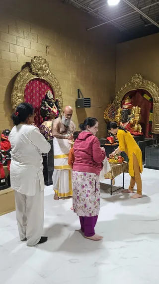 Photograph inside a temple room with gold-painted walls and two gilded arched niches holding dark deity murtis, one wearing a red-and-gold cloth and flower garland. A bald, bearded priest in a white dhoti with gold borders and bare chest stands at center, holding a plate, with rudraksha beads visible. A woman in a pink hooded top and floral skirt stands in front of him with a child beside her, while a woman in a yellow kurta and mustard leggings arranges fruit on a tray at a side table. A woman in white kurta and pants stands at left watching. Marble floor, bare feet, offerings of fruit and flowers on a table draped in gold cloth.
