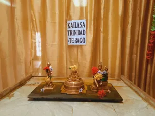A small shrine set against gold silk curtains with a white paper sign reading KAILASA TRINIDAD & TOBAGO taped to the backdrop. On a dark green stone slab sit three items: a small bronze deity figure on the left, a copper-coloured stepped sri yantra-style meru on the centre, and a bronze seated deity figure on the right, each decorated with red and yellow flowers and blue blossoms. No people are visible; the floor is pale marble with a red flower on the slab's edge.