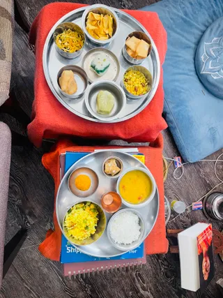 Overhead photo of two stainless steel thali plates resting on red cloth-covered seats on a dark wood floor. The upper thali holds small steel bowls of yellow and orange snacks, fried crisps, a white and green sweet, a yellow rice dish with nuts, and a pale green sweet. The lower thali holds yellow rice with peas and corn, white steamed rice, yellow dal, a small orange sweet, a copper cup, and a small piece of sweet. A blue cushion with a lotus motif sits at right beside a book with a red cover and a bottle, and the plates sit on blue notebooks and papers.