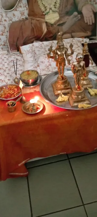Indoor altar photo for evening puja. A metal tray holds bronze deity murtis on pedestals, decorated with flower petals and yellow offerings, with a central four-armed standing deity murti. Beside the tray are a steel bowl of green-yellow prasadam with a flower on top, a bowl of orange flower petals, a small cup with a spoon, a brass oil lamp with a lit flame, a plate with petals and a burning wick, and incense sticks in a holder. The table is draped in orange-red cloth. Behind it, a large printed picture of a seated figure in saffron-brown robes with a garland, on a white patterned cloth. No people are visible.