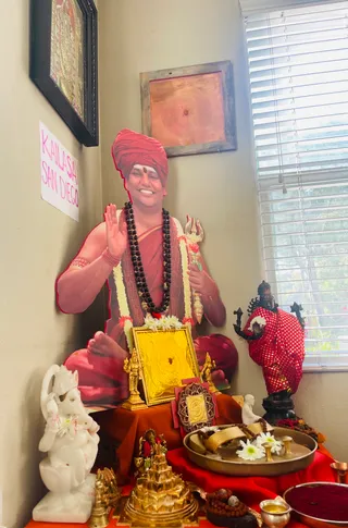 A photograph of a home shrine corner with a life-size cutout of a smiling man seated cross-legged against the wall. The cutout wears a red-orange turban with a vermilion mark on the forehead, a dark rudraksha mala and white flower garland, and raises his right hand in blessing while holding a trident. A handwritten paper sign reading "KAILASA SAN DIEGO" hangs on the left wall. In front stand a white marble Ganesha, a gold framed yantra, a gold pyramid-shaped sri chakra, small brass deities, a brass tray with white flowers and rudraksha malas, a bowl of red kumkum, and a black metal Shiva figure in a red polka-dot cloth. No physical people are present.