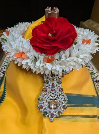 Close-up photograph of a deity murti's head and shoulders draped in a yellow silk cloth with a dark green border. A large red flower sits at the top, encircled by a garland of white blossoms with orange accents. A jeweled gold pendant set with white, green and pink stones hangs from the garland, with a gold flower or diamond-like centerpiece. The upper portion of a metal crown or headdress is visible above. A white and gold embroidered cloth lies beneath.