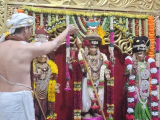 A shirtless priest in a white dhoti and white head-cloth performs alankaram and ritual worship before three deity murtis in a decorated shrine. The central golden-hued deity wears a tall gold crown with a teal and yellow cloth, a rudraksha-style necklace, a white dhoti and holds a trident-like staff; flanking figures wear embellished garlands, jewelry and silver-patterned adornments. The shrine is framed by garlands of yellow marigolds, red roses, white jasmine and pink flowers against a red backdrop and gold carved panels. The priest holds a lamp near the figures, with white sacred ash marks on his arms and chest.