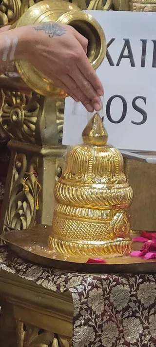 Close photo of a gold-plated Shiva lingam on a brass tray, its tiered, ridged surface shining, with pink rose petals scattered at its base. A hand with a tattoo and white sacred markings on the wrist pours liquid from a gold vessel onto the top, an abhishekam offering. Behind it, a white sign shows large black capital letters reading KAIL and LOS. Gold carved ornamentation and a maroon brocade cloth with gold floral patterns frame the altar.