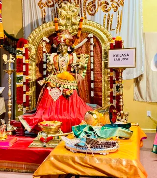 A golden deity murti is seated on a throne within a gilded arch, draped in red and orange silk and adorned with garlands of orange marigolds and white and red flowers. The figure holds a trident-like staff and wears a jeweled necklace and a red-and-gold headdress. In front, a brass pot and offering trays with a green silk cloth, ceremonial items, and a brass lamp stand are arranged on an orange cloth. A framed placard on the right reads KAILASA SAN JOSE. No people are visible in the scene.