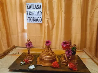 A photograph of a small shrine set on a dark wooden tray against a gold silk curtain on a marble floor. A printed white paper sign taped to the curtain reads KAILASA TRINIDAD & TOBAGO. In the centre stands a tiered copper-toned shivalinga-style peetha with a stepped base, topped with a pink hibiscus flower. To the left is a small copper-toned statue of a standing male figure with a pink flower draped over his shoulder. To the right is a copper-toned deity murti seated in a crowned form beside a smaller figure, decorated with pink and orange flowers and a green cloth. Scattered red and white flowers lie on the tray in front. No people are visible.