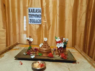 A small shrine set against gold silk drapes. A paper sign reading KAILASA TRINIDAD & TOBAGO is taped to the drapery. On a dark wooden board sit copper-toned deity murtis and a tiered copper shivalinga-like structure, decorated with white plumeria flowers, red ixora blossoms and green leaves. A small steel bowl holds white offerings with red flowers scattered nearby. No people are visible.