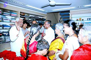 A photograph inside a clothing shop with shelves stacked with folded fabrics in blues, purples and reds, and a glass display case on the right holding books. An older man in a white shirt and white dhoti, with white hair, gestures with one raised hand while speaking to a group. Around him stand several women in saris: one in red with a long side braid, one in a yellow-and-maroon sari with grey hair, another in white with a black beaded necklace, and an older woman in a red sari at the right edge. A young boy stands in the lower foreground beside red flower garlands and petals on a table. A man in a striped shirt and another in a white shirt stand behind. A ceiling fan and fluorescent lights are overhead. The man in white is not identifiable as a specific person from appearance here, so no a person identification is made.