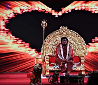 A man sits on an ornate gilded throne with carved floral and scroll details and red cushions, on a red stage in front of a large backdrop of radiant red and orange light rays. He wears a reddish-brown robe, a white flower garland, and a red tilak on his forehead, and his hands are folded in his lap. A tall silver trident stands beside the throne, and a row of bronze lion statues sits at the base of the stage. A small wooden side table holds a cup. Swamiji (probable): the seated man has long dark hair and a full beard, and his robe, garland and throne mark him as a central figure on stage, though this is an appearance-based label only.