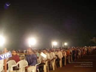 Night photo of a large outdoor satsang audience seated in rows of white plastic chairs behind a rope barrier, lit by bright floodlights on poles. Foreground men in blue and white shirts and a woman in a red sari sit near a blue and white VIP sign on a wooden stake. Many more people in the rows behind, some in orange and maroon clothing, face toward the unseen stage. No clearly identifiable a person is visible among the seated audience.