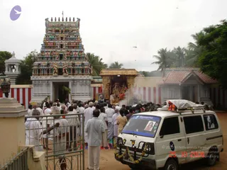 Outdoor photograph of a South Indian temple procession at a colourful gopuram with tiers of painted deity sculptures. A crowd of devotees, mostly men in white dhotis and shirts with some women in saris, stands gathered in front of a gilded palanquin or shrine carrying two decorated deity murtis with garlands. Smoke rises from the front of the shrine. A white van decorated with a garland and a portrait is parked in the foreground beside a metal gate. Palm trees and a red-tiled shrine structure stand behind. A portrait printed on the van reads Nithyanandam. Dozens of people are visible.