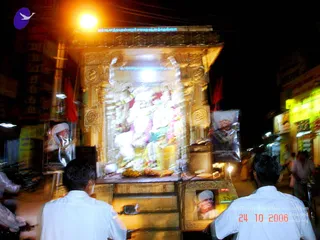 Night photo of a decorated temple-style float, a ratha, on a street in Tamil Nadu during a procession. The gilded carved structure is hung with garlands in white, pink and green and holds a deity murti at its centre, lit by a bright lamp. Framed portraits of a saffron-robed guru wearing a white head-wrap are mounted on the float's sides. Two men in white shirts stand at the front edge with their backs to the camera, facing the float. Red flags and shopfronts with lit signs line the street, and the image is motion-blurred.