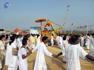 A festival procession on a dusty road with a flower-decorated float or chariot in the centre. The float is heavily garlanded with yellow and orange marigold strands and has an orange canopy umbrella and tassels. A figure seated on the float wears orange and red clothing and is seen from a distance. Around it, many people in white ritual dress walk in lines, several holding hands in a chain, with yellow and orange scarves and orange-flag-carrying participants behind. Orange flags fly on poles, and a roadside banner with a portrait and printed text stands near a chain-link fence under a clear pale sky.