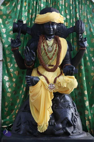 A black stone statue of a multi-armed deity seated on a rocky pedestal, draped in yellow fabric with a yellow turban and a red tilak on the forehead. It wears layered rudraksha malas and a jewelled pendant, and its raised hands hold a trident-like staff and a drum-shaped object. Behind it hangs green cloth with gold floral print. Carved reliefs of animals and figures decorate the base. No living people are visible.
