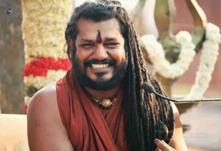 Close-up photograph of a smiling bearded man with long dreadlocked hair falling over his shoulders, wearing a rust-red sleeveless wrap and a gold pendant on a chain, with a dark vertical tilak mark on his forehead. A microphone on a stand is visible at right, and behind him are white jasmine garlands with red flower strands and a brass ritual vessel. a person: long dreadlocked hair, rust-red wrap and ornate gold pendant with a microphone at a satsang, matching the page's note of a live talk.