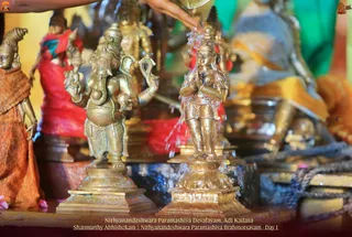 Photo of a ritual abhishekam in progress over brass deity murtis. A hand at the top edge pours water from a vessel onto a small standing brass figure at centre-right, water streaming down its body. Beside it stands a brass Ganesha murti with elephant head, raised arms holding a modak and an axe-like implement, on a stepped base. Other brass and gilded murtis, some draped in red and orange cloth, crowd the frame. Caption at bottom reads Nithyanandeshwara Paramashiva Devalayam, Adi Kailasa and Shatmurthy Abhishekam, Nithyanandeshwara Paramashiva Brahmotsavam - Day 1.