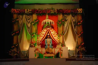 A photograph of an ornate stage set for a satsang. A seated figure sits on a large gold throne with red cushions, framed by a golden arched backdrop decorated with red, yellow and purple garlands. A golden canopy with hanging beads crowns the throne. Above, a long garland of red and yellow flowers hangs across the top of the stage, with green foliage and floral swags on grey and gold draped curtains behind. The seated figure wears a turquoise-blue kurta with a light gold shawl and a small white headpiece. Flower arrangements stand at the stage base, and the scene is lit in green and warm tones.