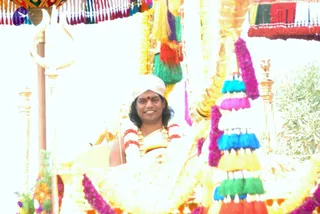 A smiling man sits in an ornate golden palanquin or ratha decorated with colourful tassels in blue, green, yellow, red and pink, and fresh magenta and yellow flower garlands. He wears a white turban with a gold-toned band and a red bindi, a white shawl over bare shoulders, and a layered garland of red and white flowers with a gold necklace. Golden finials, a trident-like staff and a green plant appear at the sides. The scene is bright and outdoors.