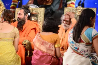 A photograph of a seated group on a stage at an indoor event. Two elderly-looking men sit on gold-framed chairs with lion-head finials, both in orange-saffron robes. The man on the right has white hair and a long white beard, touching his head with one hand, with a red mark on his forehead. The man on the left has long dark hair, a full beard and a white earpiece, wearing an orange robe. Several women sit in the foreground with their backs to the camera, wearing a yellow salwar, an orange blouse with a pink-and-gold sari, and a white sari with colourful embroidery. A gold lion figure sits in the foreground. Behind them is a blue backdrop with a decorated statue.